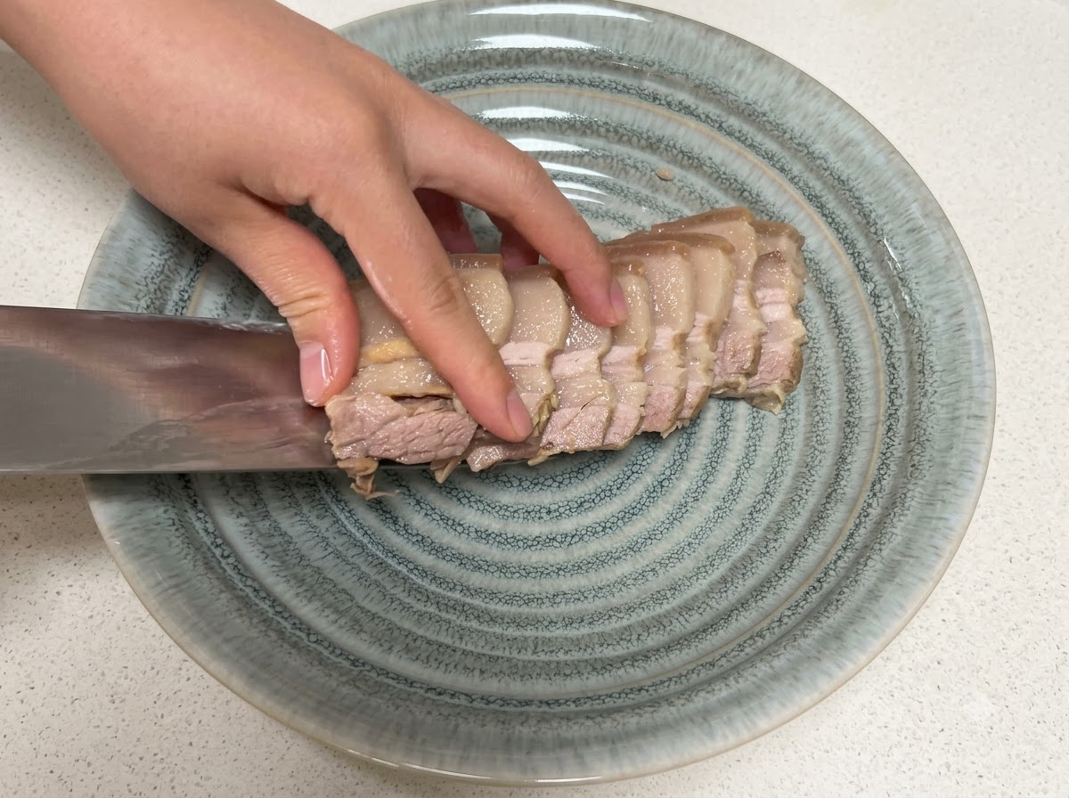 Rebanadas de panceta de cerdo cocida siendo transferidas a un plato gris texturizado con un cuchillo.