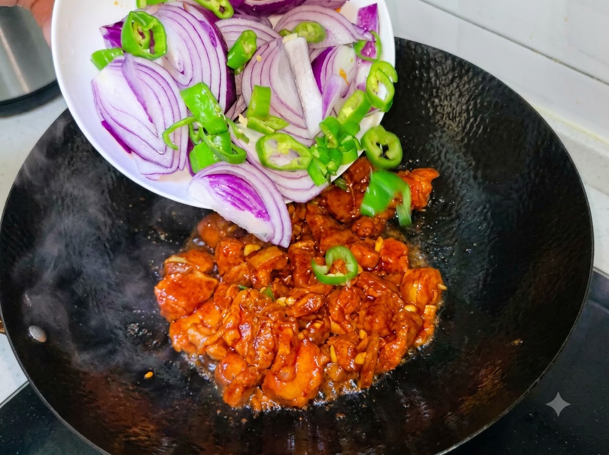 Rodajas de cebolla morada y chiles verdes vertiéndose desde un plato blanco a un wok con panceta de cerdo en cocción.