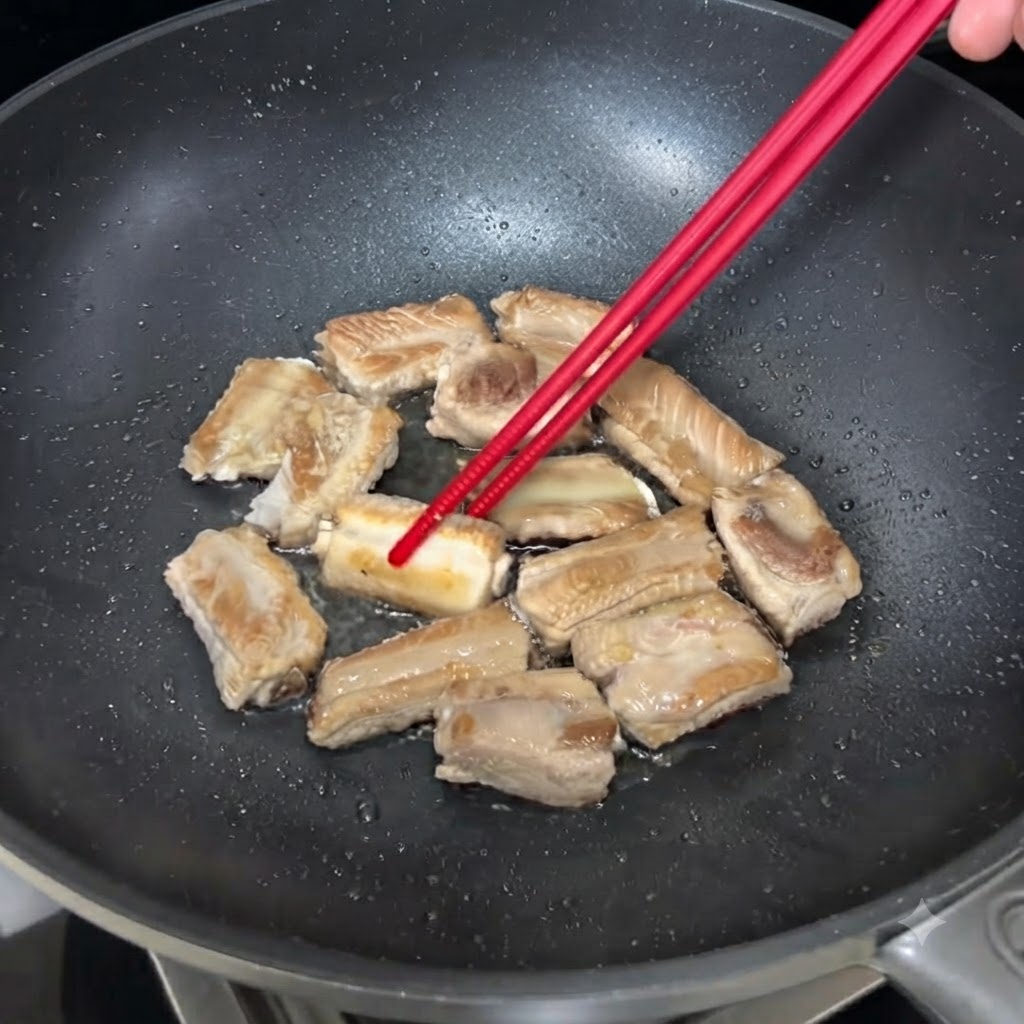 Costillas de cerdo chisporroteando en una sartén y siendo volteadas con palillos para que se doren de manera uniforme.