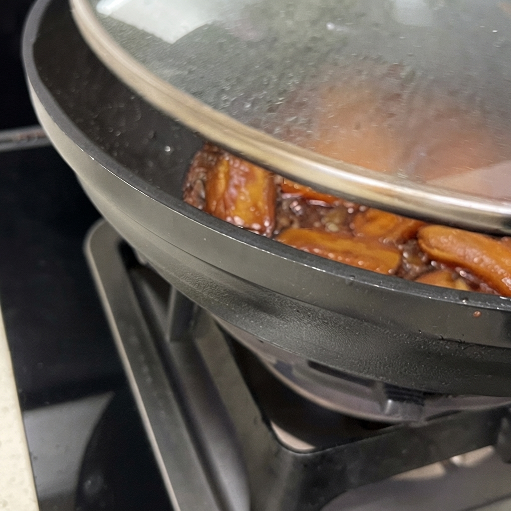 Primer plano de un wok con la tapa parcialmente levantada, mostrando costillas de cerdo cocinándose a fuego lento en una salsa marrón espesa y brillante.