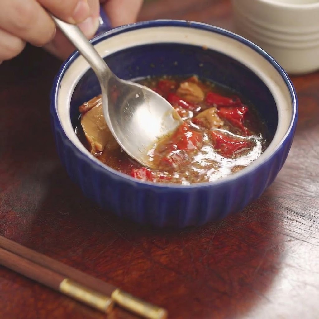 Una cuchara triturando y mezclando tofu rojo fermentado y condimentos dentro de un pequeño bol de cerámica azul.