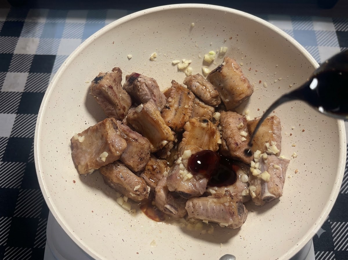 Un pequeño chorrito de salsa de soja oscura siendo vertido en la sartén con costillas de cerdo fritas y ajo picado.
