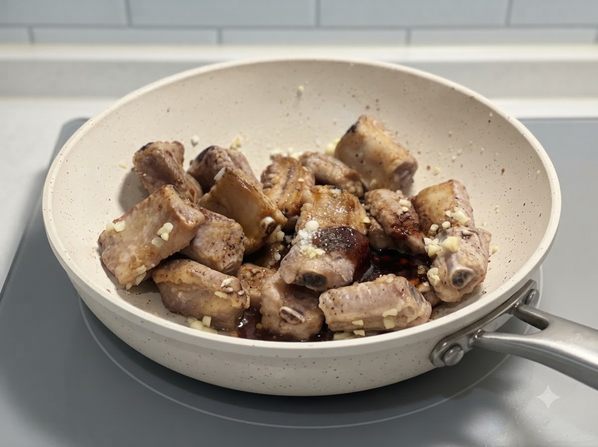 Costillas de cerdo siendo mezcladas junto con condimentos y ajo picado en una sartén blanca sobre una estufa.