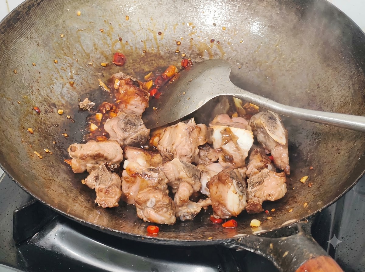 Costillas de cerdo fritas siendo mezcladas en un wok con salsa, chiles picados y ajo usando una espátula de metal.