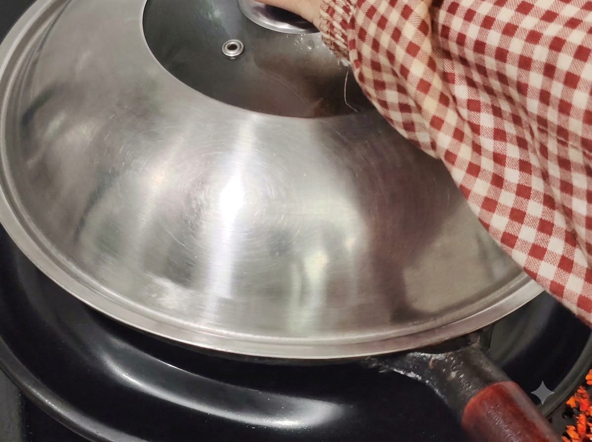 Una tapa de wok de metal cubriendo el wok en una estufa, sostenida por una mano con una manga a cuadros rojos y blancos.