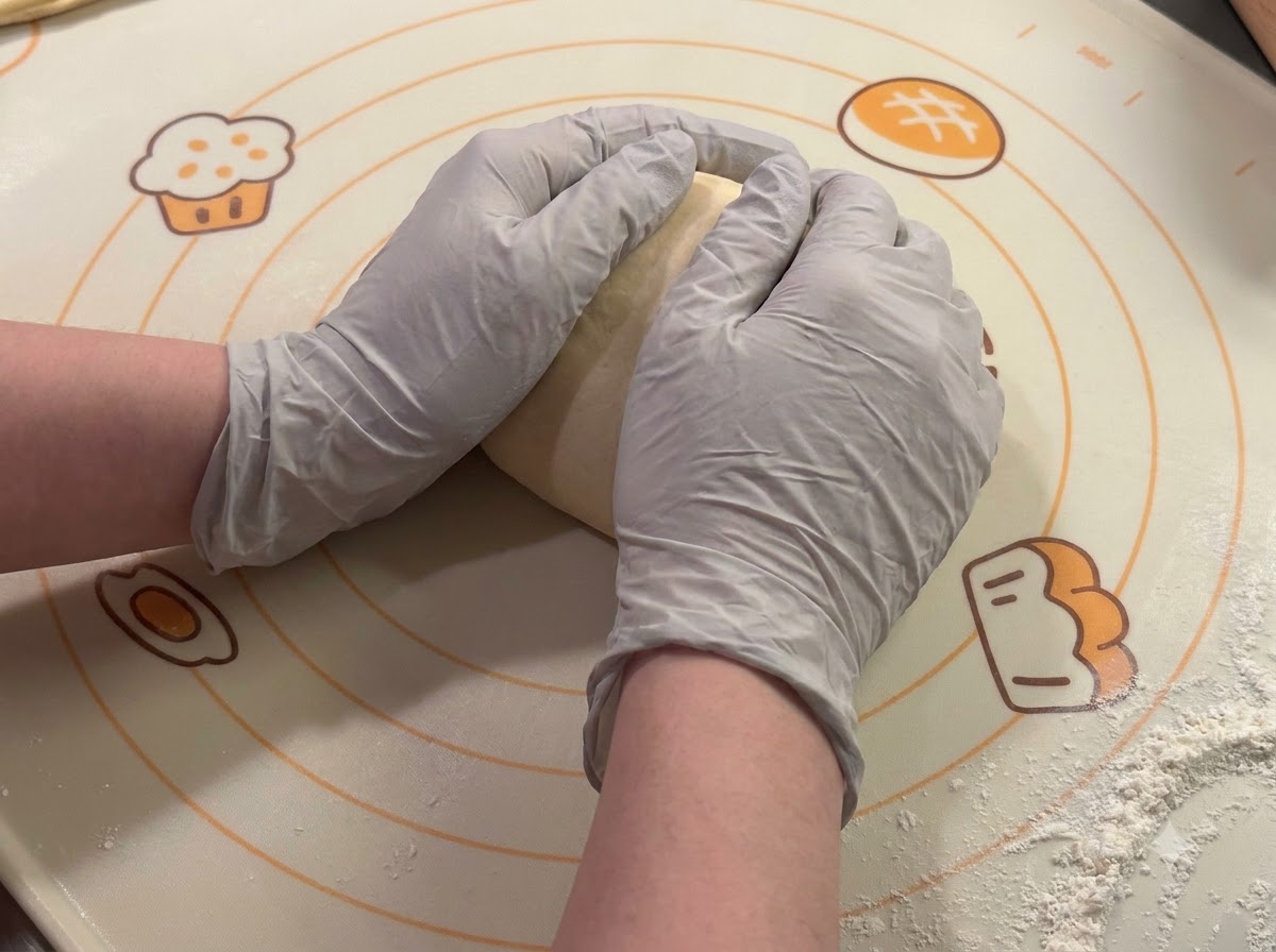Manos con guantes dando forma y redondeando porciones de masa sobre una esterilla de horno.