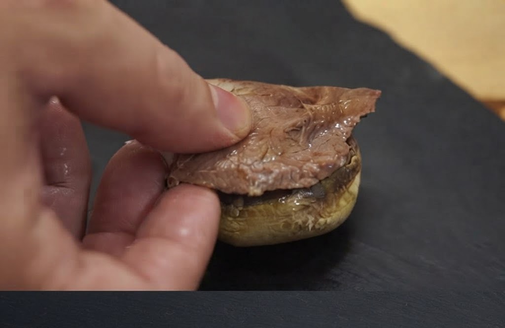 Una mano colocando un trozo de carne cocinada sobre la base de un sombrero de champiñón horneado.