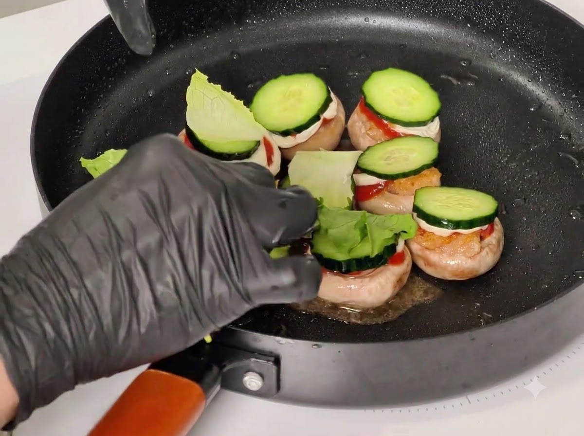 Una mano enguantada colocando palillos a través de los ingredientes apilados en las tapas de los hongos dentro de una sartén negra.