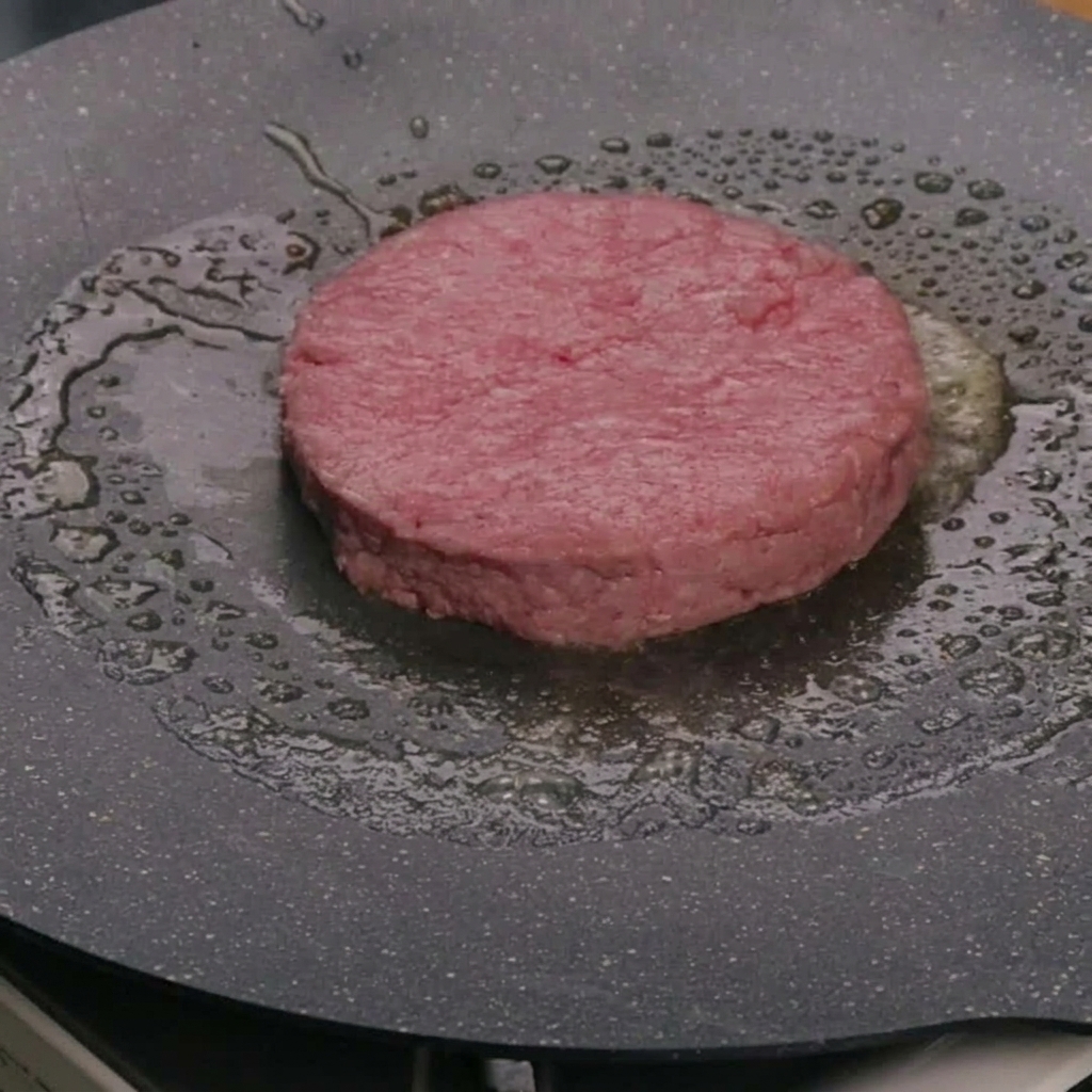 Una hamburguesa de carne cruda cociéndose en mantequilla dentro de una sartén caliente.