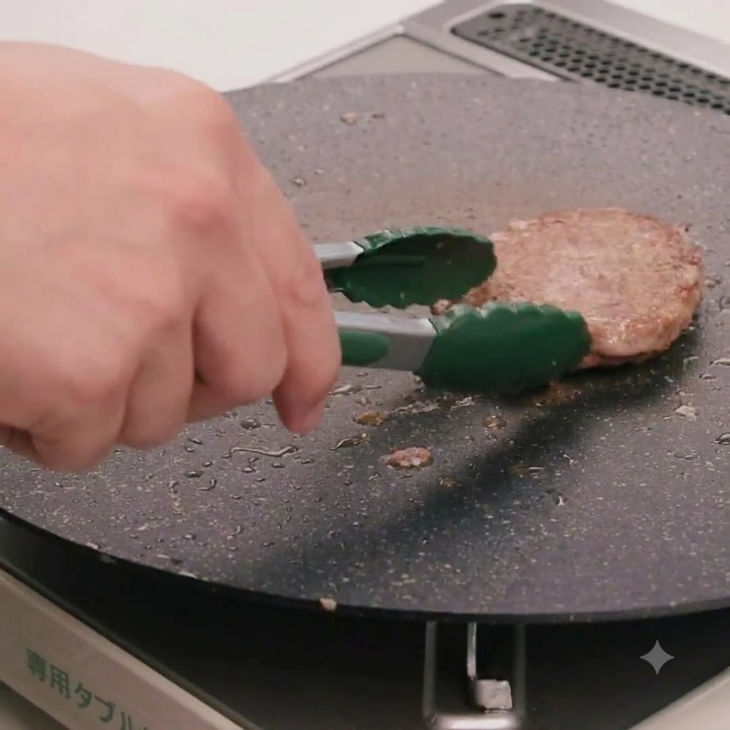 Una mano usando pinzas de cocina verdes para voltear una hamburguesa de carne en una sartén plana.