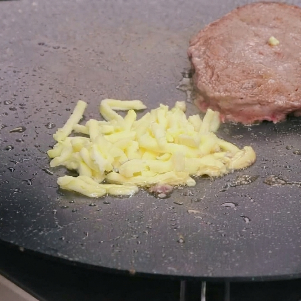 Queso mozzarella rallado derritiéndose encima de una hamburguesa de carne frita en una sartén caliente.
