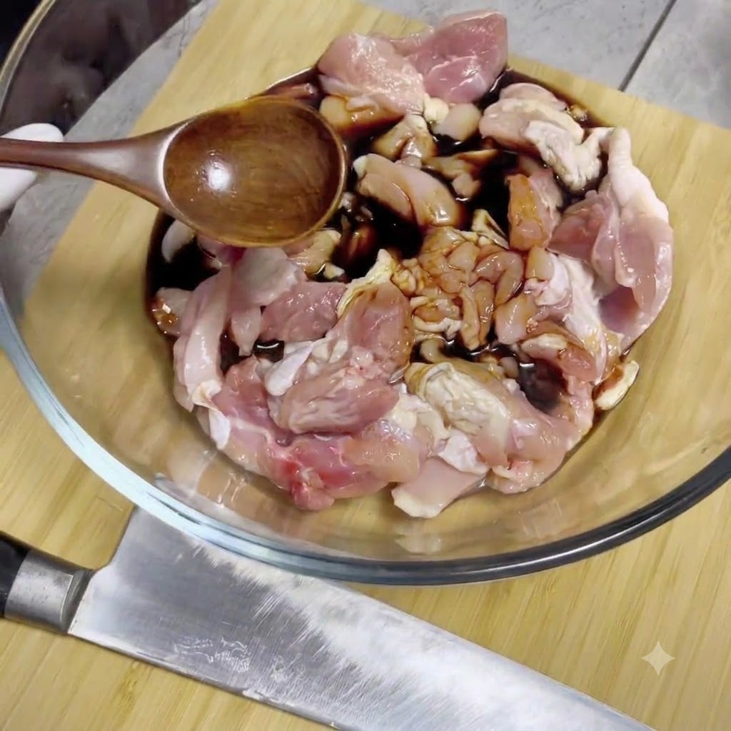 Salsa de soja siendo vertida desde una cuchara de madera sobre trozos de pollo crudo dentro de un bol de vidrio para mezclar.