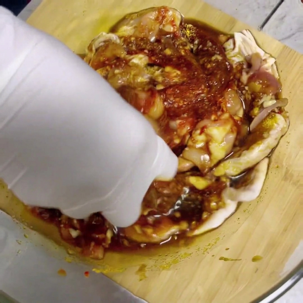 Una mano con un guante blanco mezclando trozos de pollo crudo y salsa roja para marinar en un bol de vidrio.