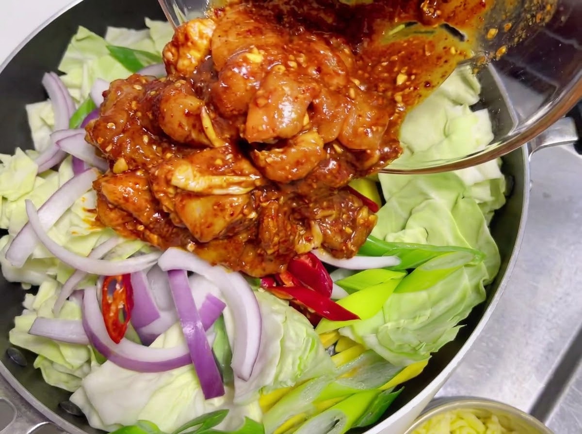 Pollo crudo marinado cubierto con una salsa roja picante siendo vertido de un bol de vidrio sobre una capa de repollo crudo y cebollas.