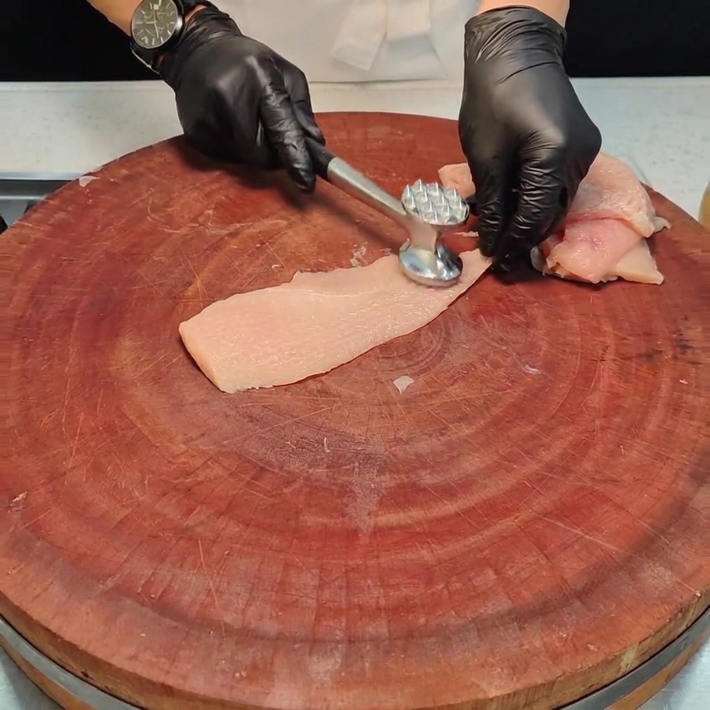Una pieza de pechuga de pollo siendo aplanada con un mazo de metal para carne sobre una tabla de madera.