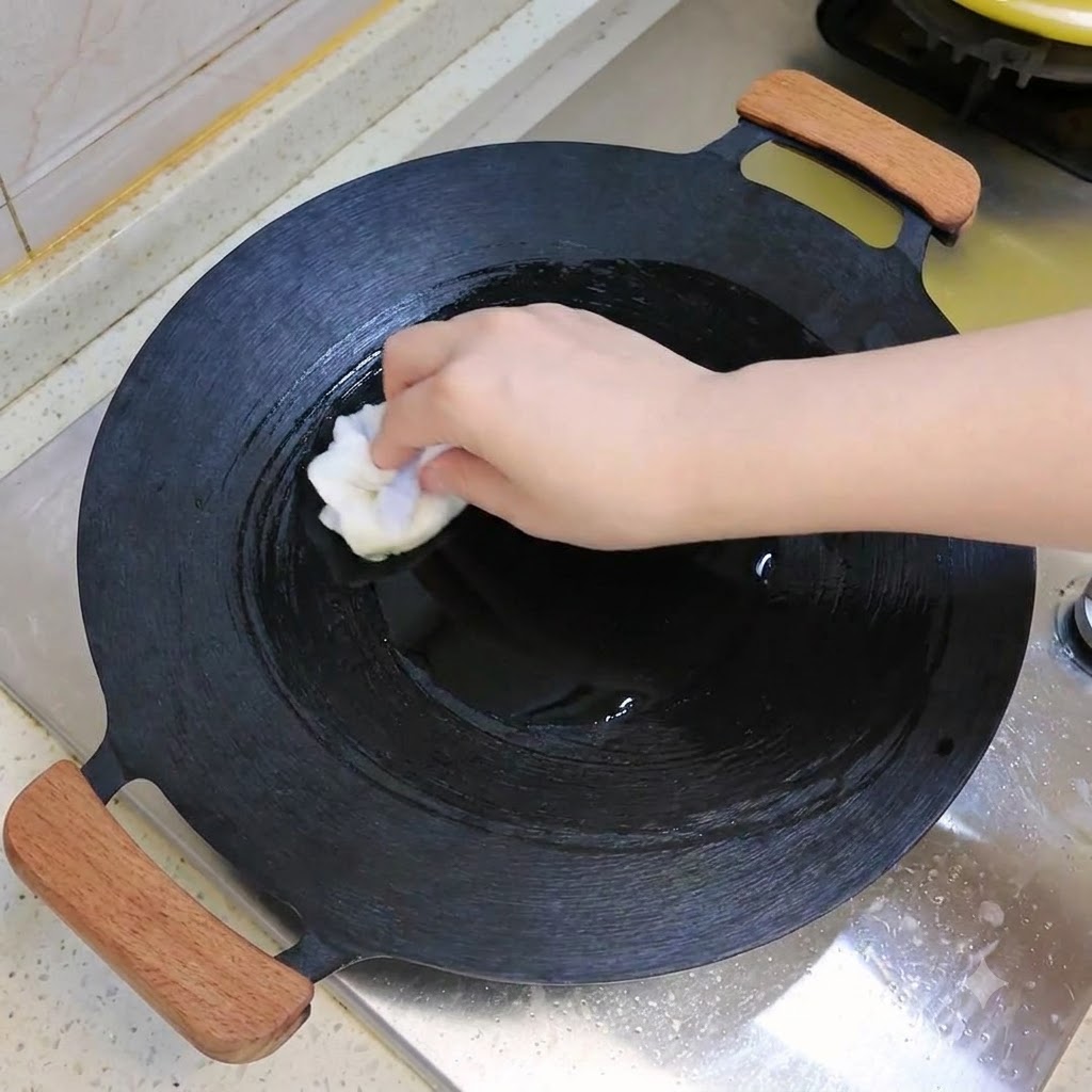 Un wok oscuro siendo limpiado o aceitado con un paño blanco sobre una estufa.