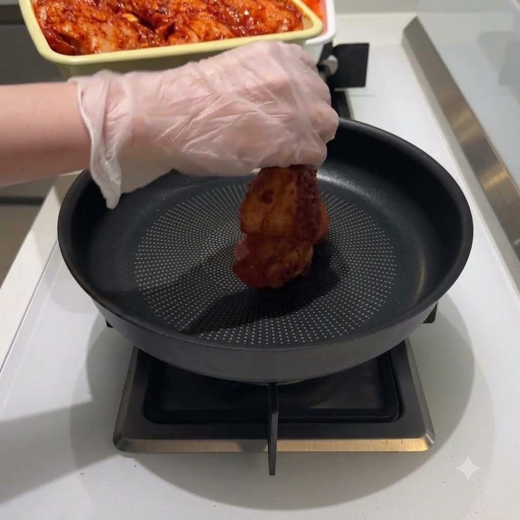 Una mano con guante colocando un trozo de pollo marinado rojo en el centro de una sartén negra.