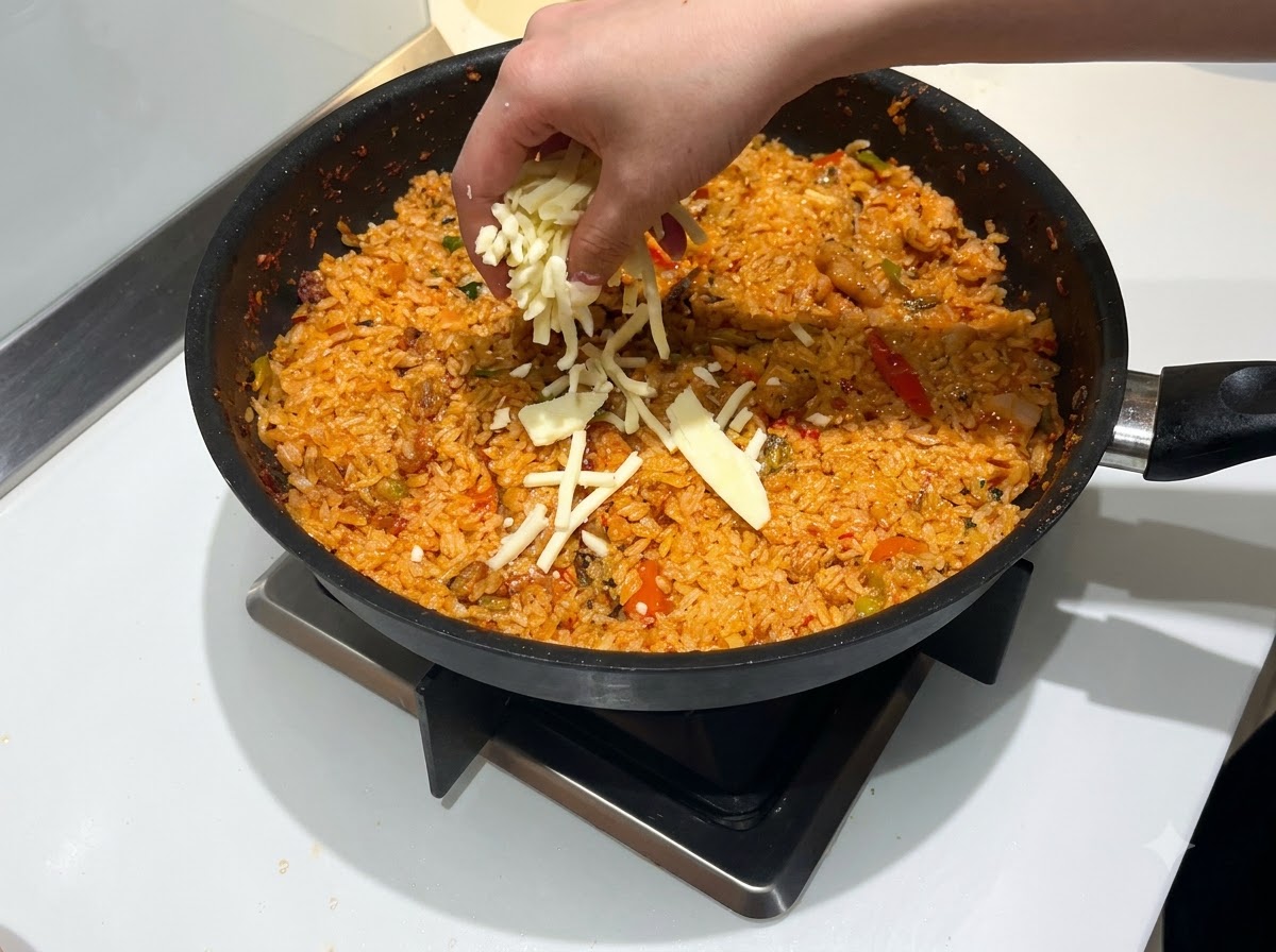 Una mano espolvoreando una generosa cantidad de queso mozzarella rallado sobre una sartén de arroz frito rojo y picante.