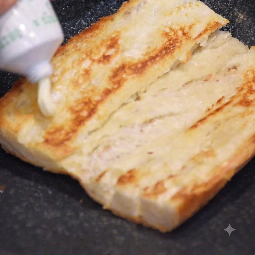 Mayonesa saliendo de un tubo sobre una mitad de baguette dorada y tostada.