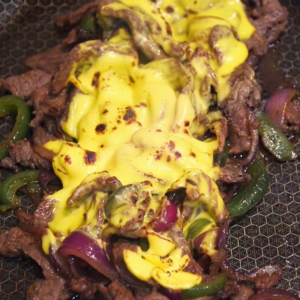 Un soplete de cocina flameando la capa de queso amarillo fundido sobre la mezcla de carne y verduras en una sartén.