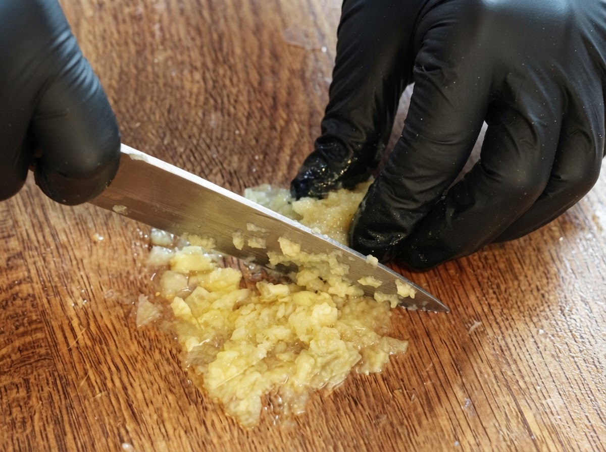 Un par de manos con guantes usando un cuchillo para picar y hacer puré la pulpa de limón sobrante en una tabla de cortar de madera.