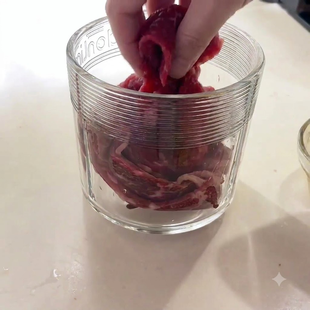 Una mano colocando trozos de carne de res cruda en el recipiente de vidrio de un procesador de alimentos.