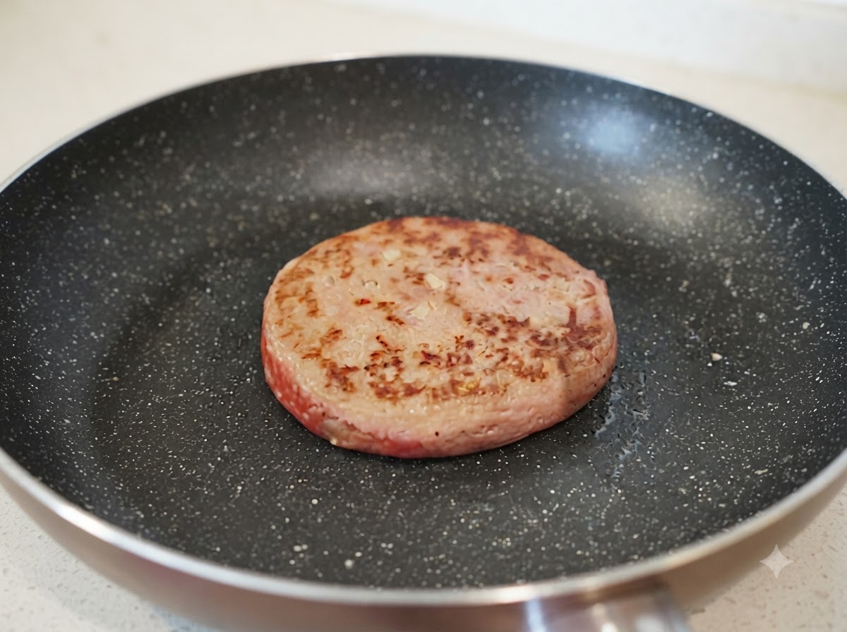 Un pequeño chorro de agua siendo vertido en una sartén junto a una hamburguesa parcialmente dorada.