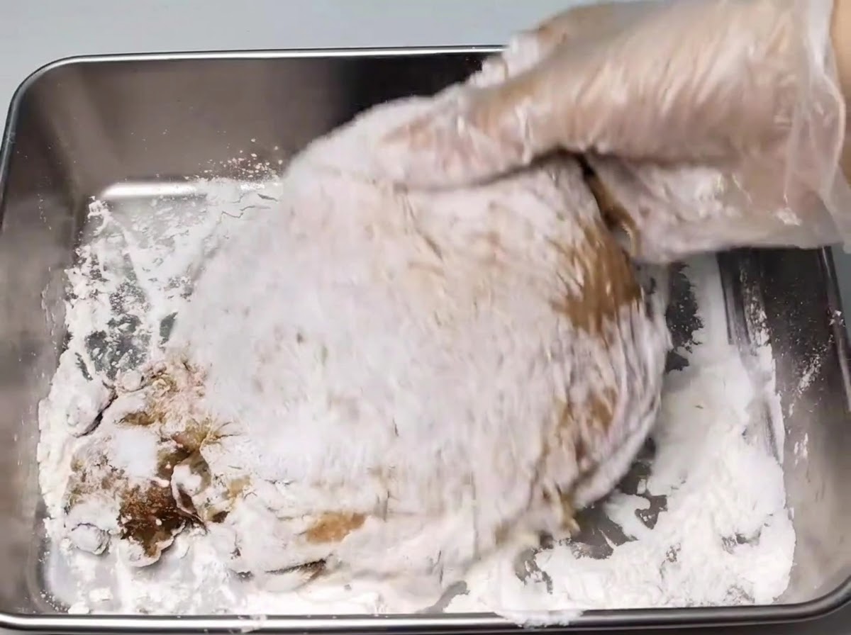 Una mano con guante cubriendo un muslo de pollo marinado con una capa de fécula blanca dentro de una bandeja de metal.