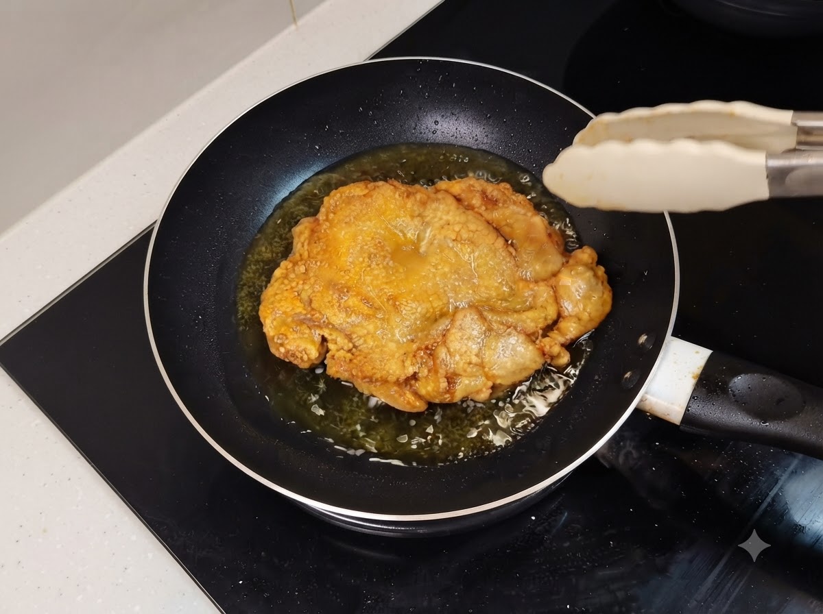 Un muslo de pollo frito de color dorado chisporroteando en una sartén aceitosa con pinzas de cocina cerca.