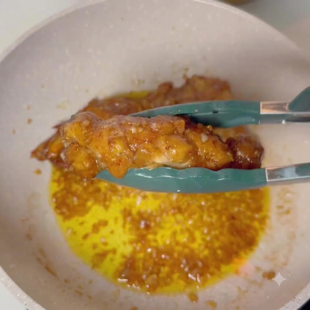 Pinzas sosteniendo un muslo de pollo glaseado sobre una sartén con salsa de mantequilla y ajo burbujeante.