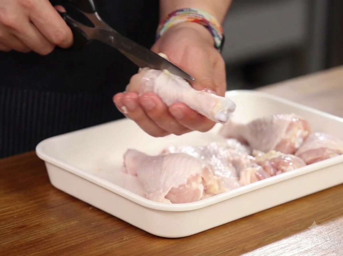 Manos usando tijeras de cocina para cortar alrededor del hueso de un muslo de pollo dentro de una bandeja blanca.
