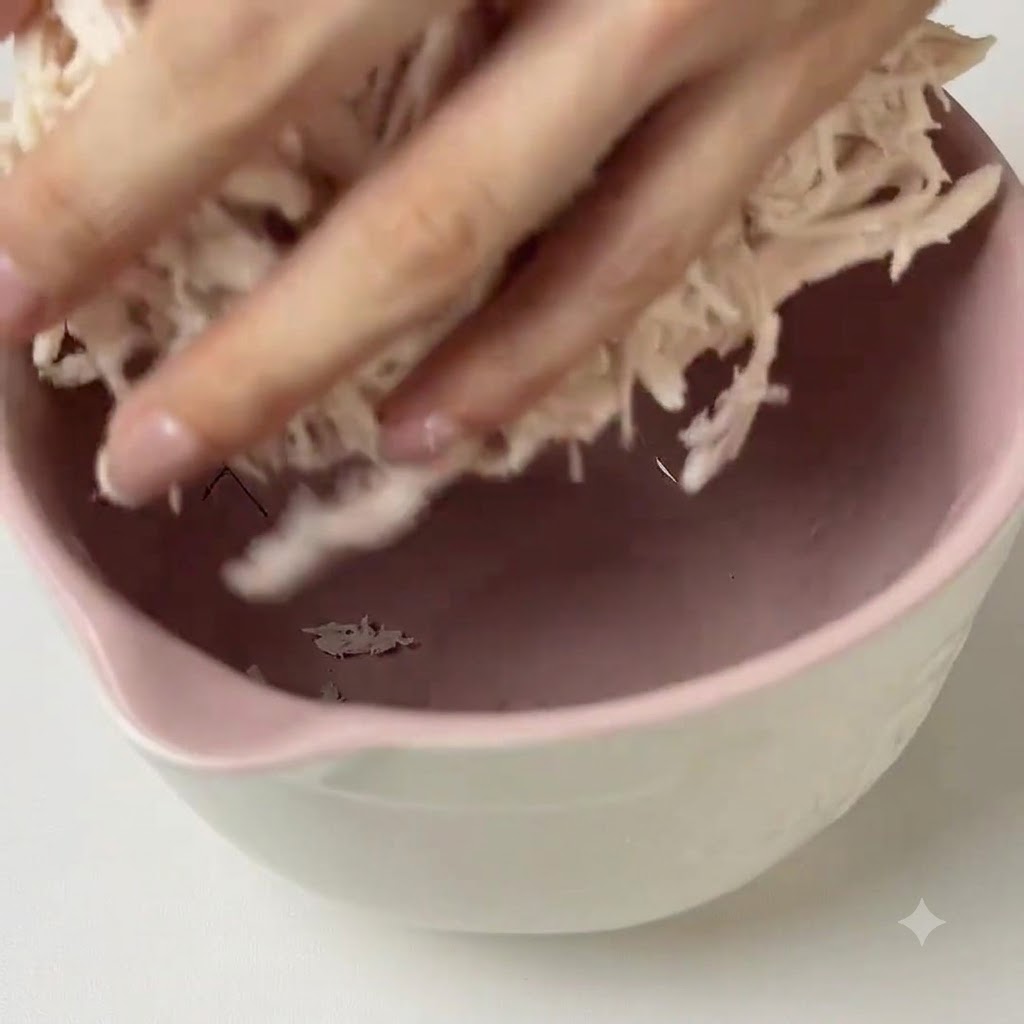 Manos mezclando salsa de pesto verde en el pollo desmenuzado dentro de un bol de mezcla rosa.