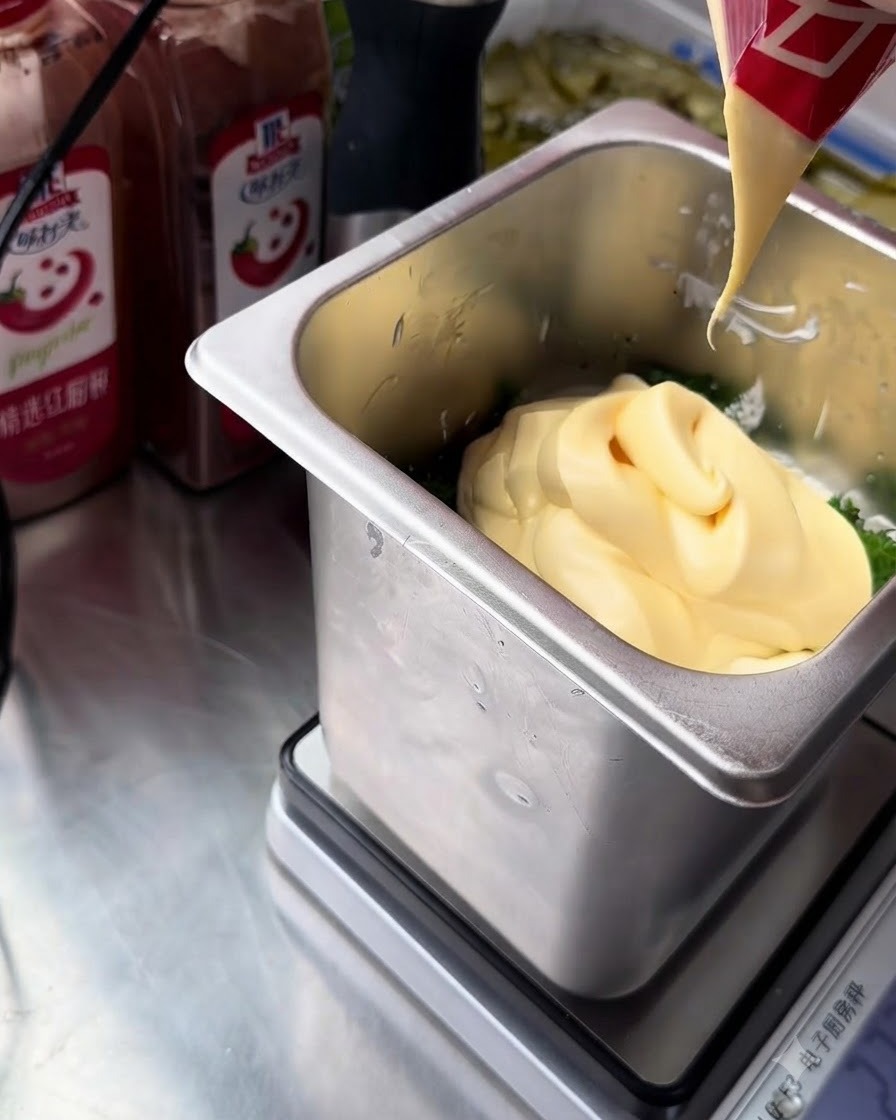 Un chorro de mayonesa de yema de huevo de color amarillo intenso siendo exprimido desde un paquete rojo y blanco en el recipiente con los ingredientes tártaros.